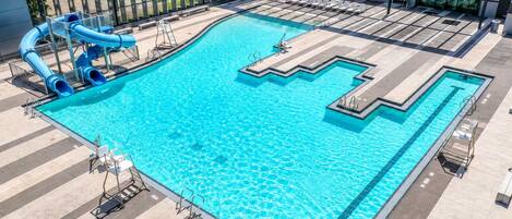 A large outdoor swimming pool with a slide and multiple lanes, surrounded by a modern building with large windows.