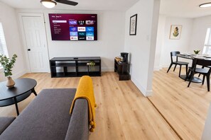 Ein modernes Wohnzimmer mit flachbildfernseher an der Wand, einer Couch, einem Couchtisch und einem Essbereich mit Tisch und Stühlen.