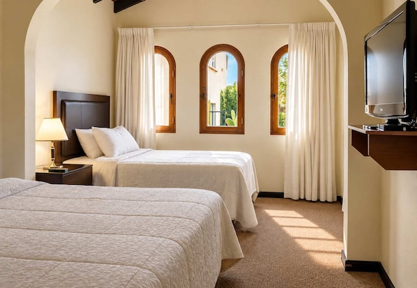 Habitación de hotel con dos camas, un televisor y tres ventanas con cortinas.