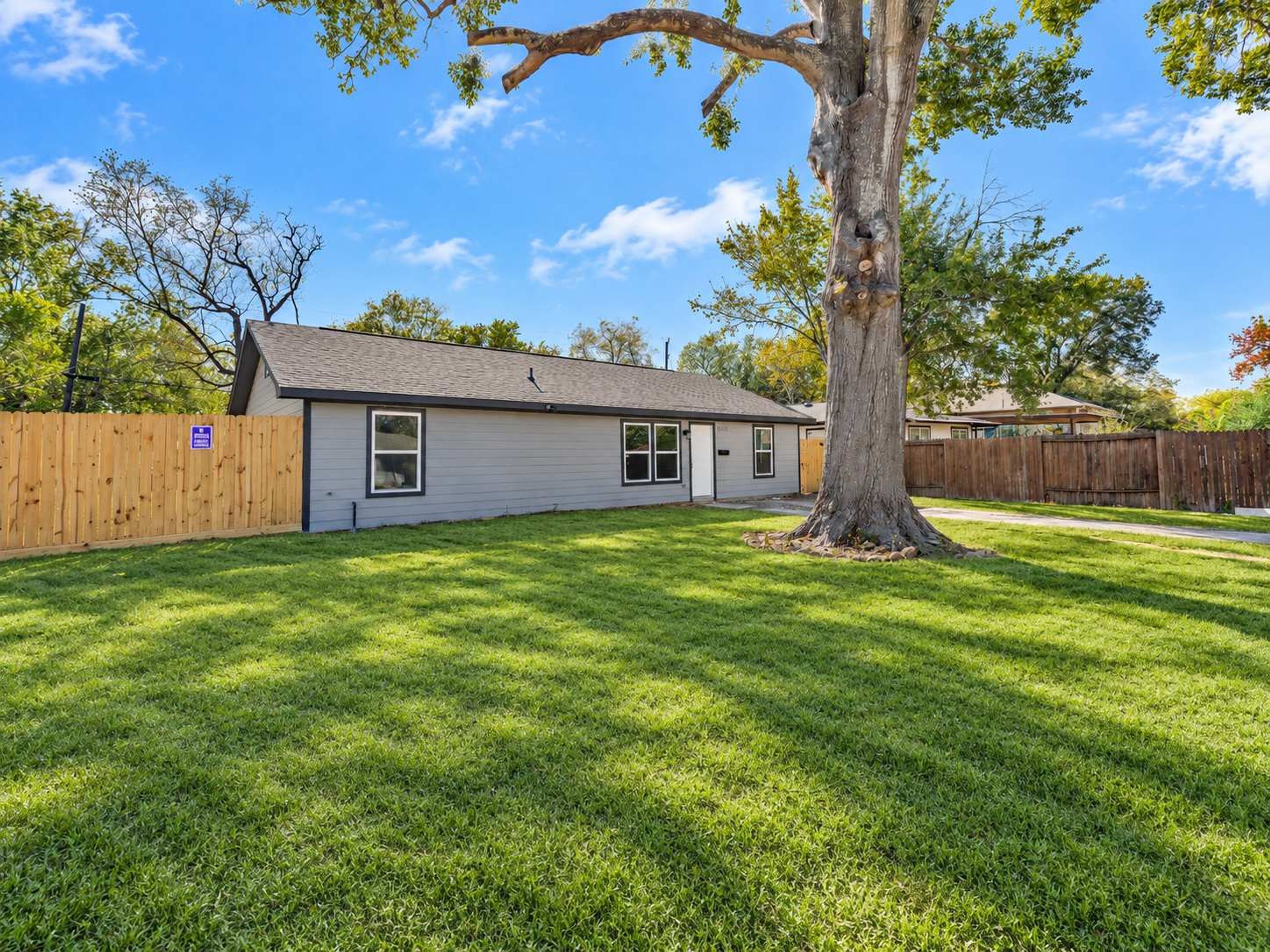 A single-story house with a fenced yard and a large tree in front.