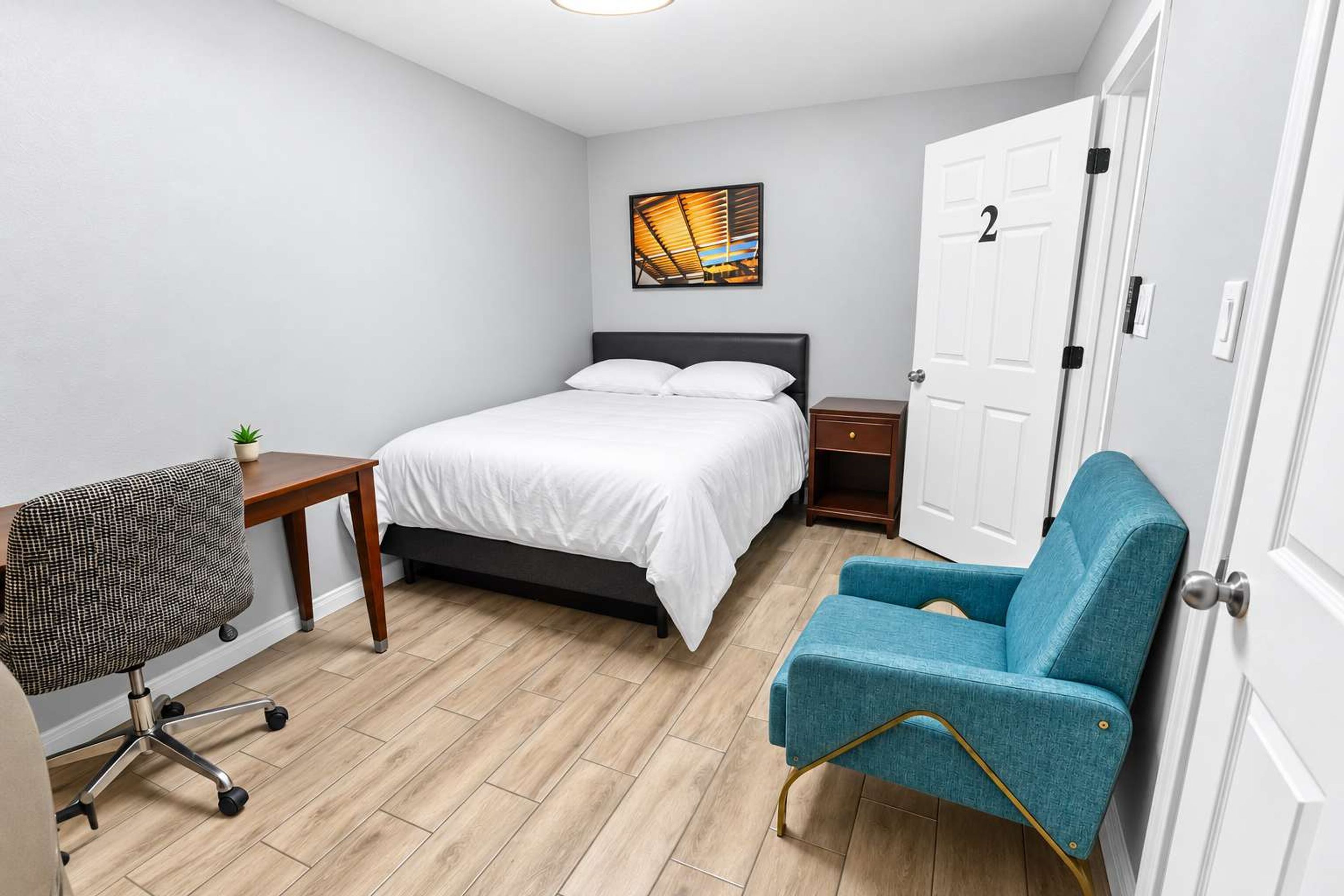 A small, neatly arranged bedroom with a bed, a desk, a chair, and a door.