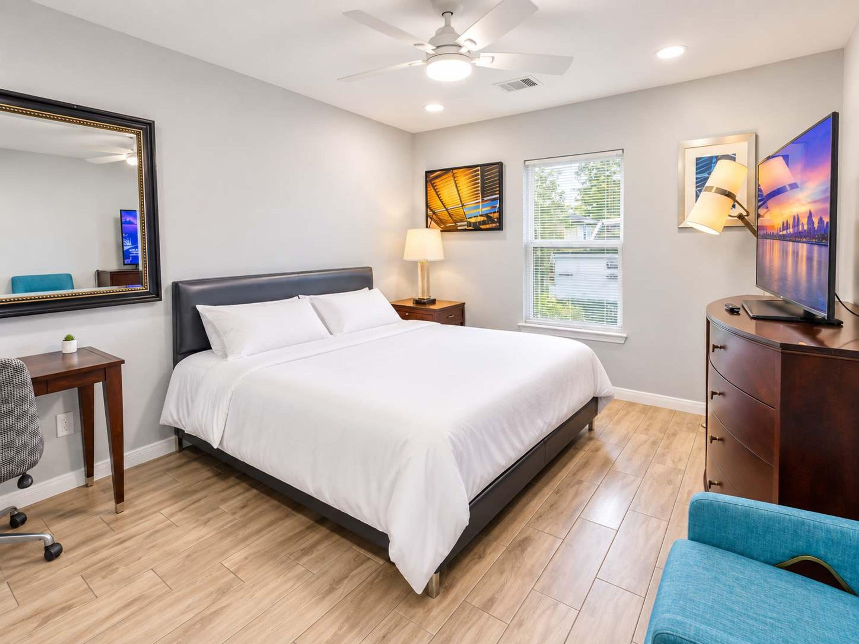 A hotel room with a bed, a desk, a chair, a dresser, a mirror, a ceiling fan, and artwork on the walls.