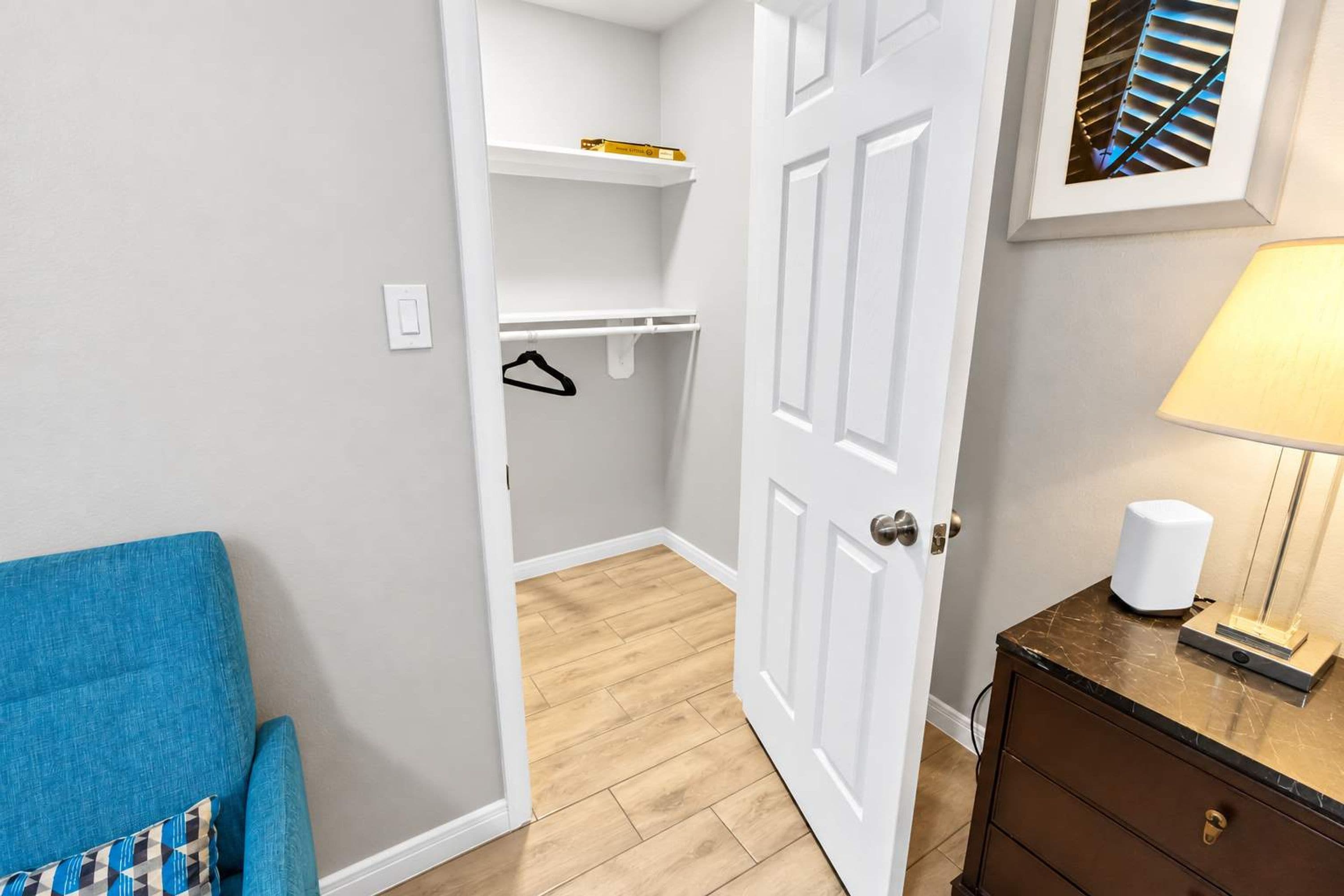 Una habitación con piso de madera, una puerta blanca, una silla azul, un aparador y una lámpara.