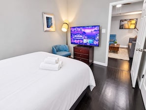 Hotellihuone, jossa on sänky, televisio, tuoli ja työpöytä, jossa on lamppu.