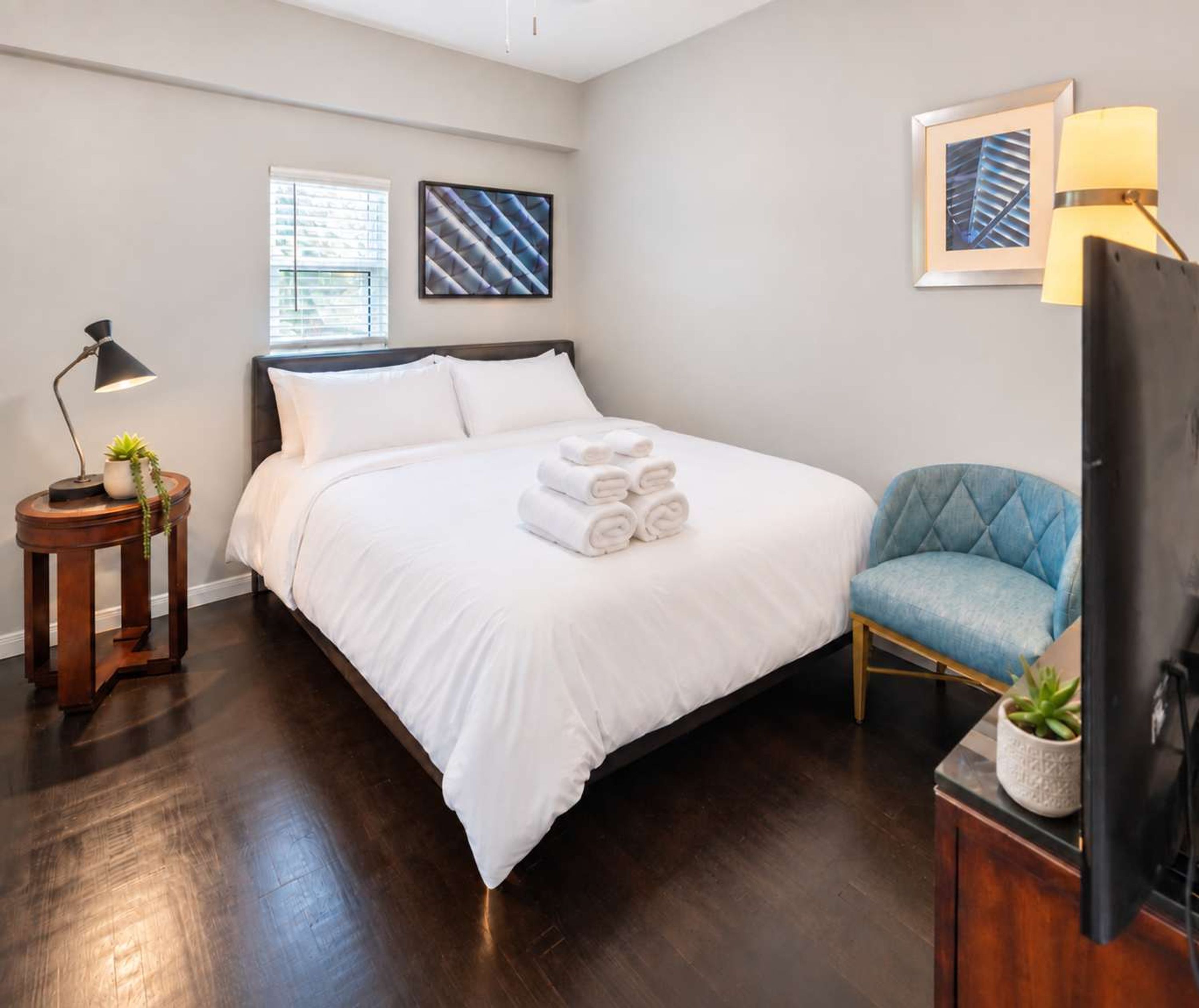 A hotel room with a bed, a side table with a lamp, a chair, and a television.