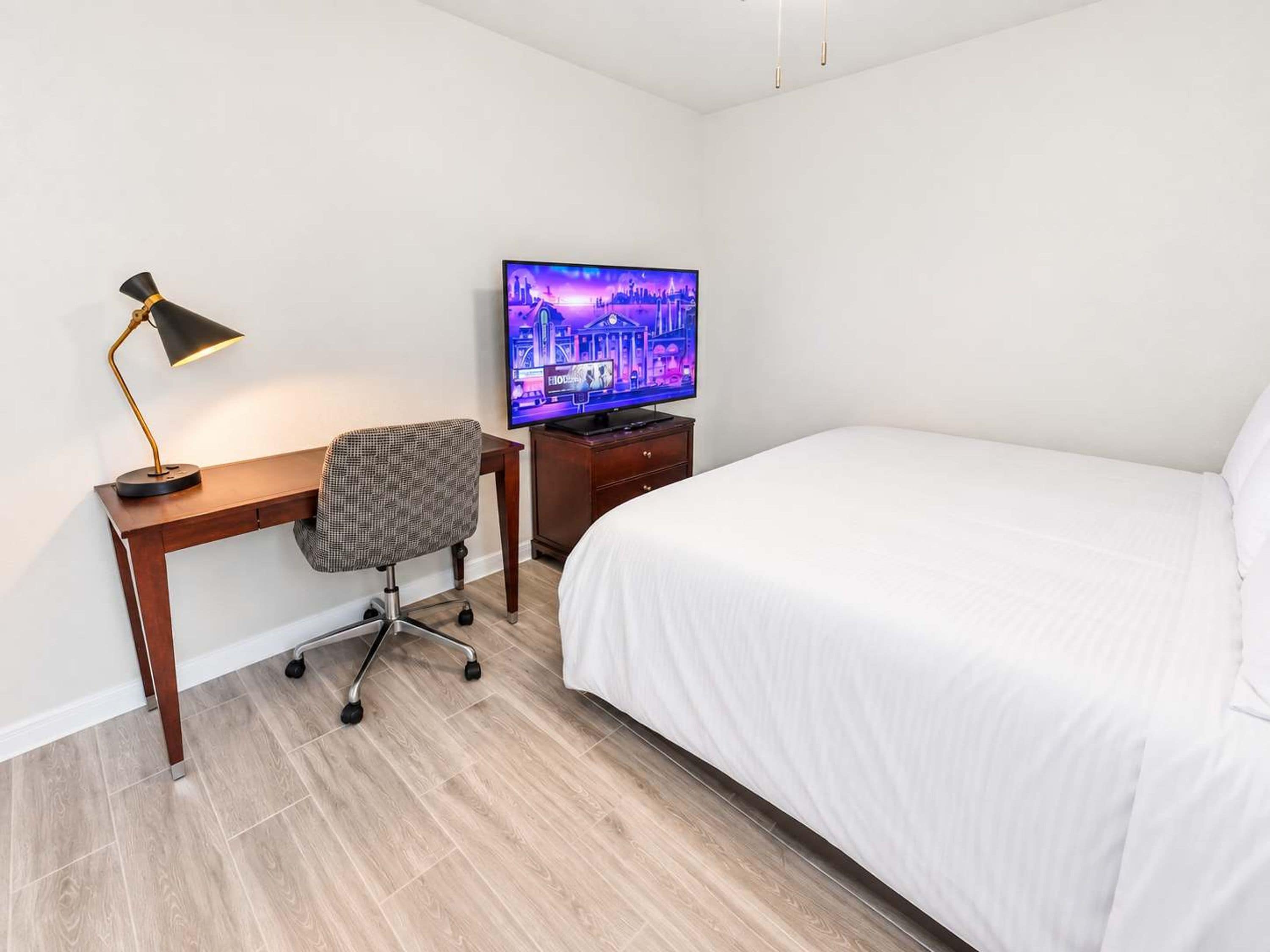 A hotel room with a bed, a desk with a chair, a television, and a desk lamp.