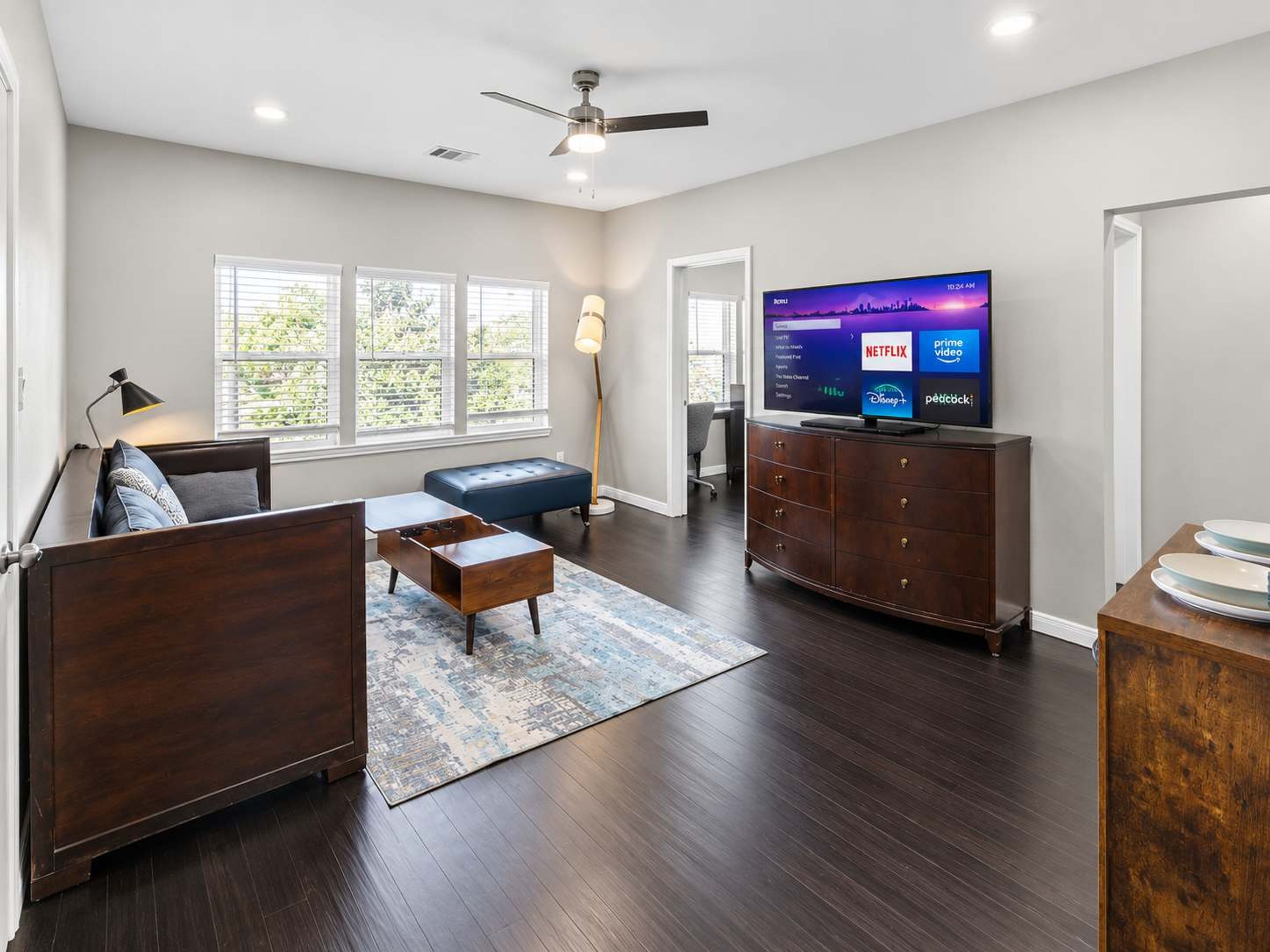 Een moderne woonkamer met een flatscreen televisie, donker houten meubels, een blauwe bank en een groot raam met zonwering.
