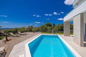 A rectangular swimming pool with clear blue water, surrounded by a paved deck, and a modern building with large windows to the right. - Villa Mate With Pool and Beautiful sea View (Krk)
