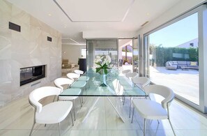 A modern dining area with a glass table and white chairs, a fireplace, and a view of the outdoors. - Villa SIA With Private Pool (Krk)
