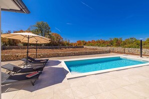 Ein Schwimmbecken mit Liegestühlen und Sonnenschirmen, umgeben von einer Steinmauer und Bäumen.