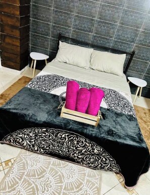 Een bed met een zwart-wit beddeken, twee witte nachtkastjes en een houten doos met roze kussens.