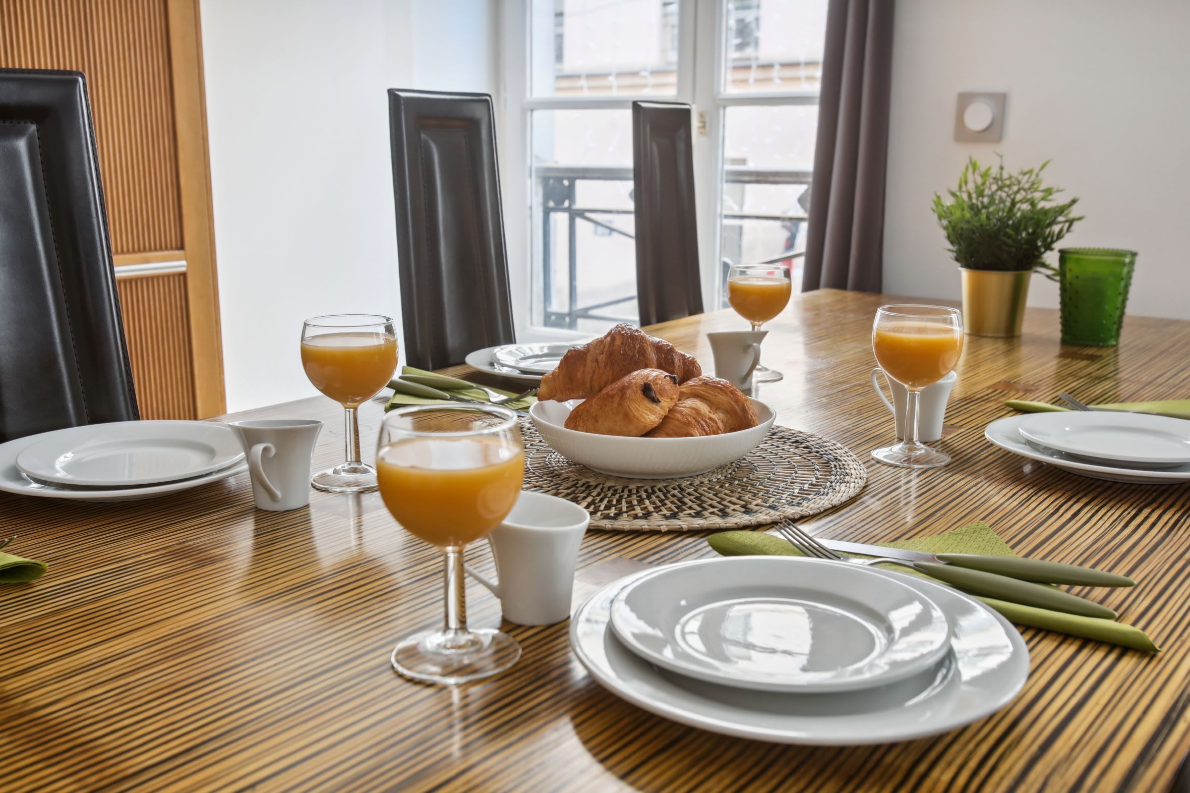 Une table en bois dressée pour le petit-déjeuner, avec des croissants, du jus d’orange et une plante verte.