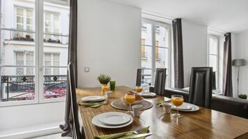 Een eetgedeelte met een houten tafel gedekt voor het ontbijt, grote ramen met gordijnen en uitzicht op een balkon met bloemen.