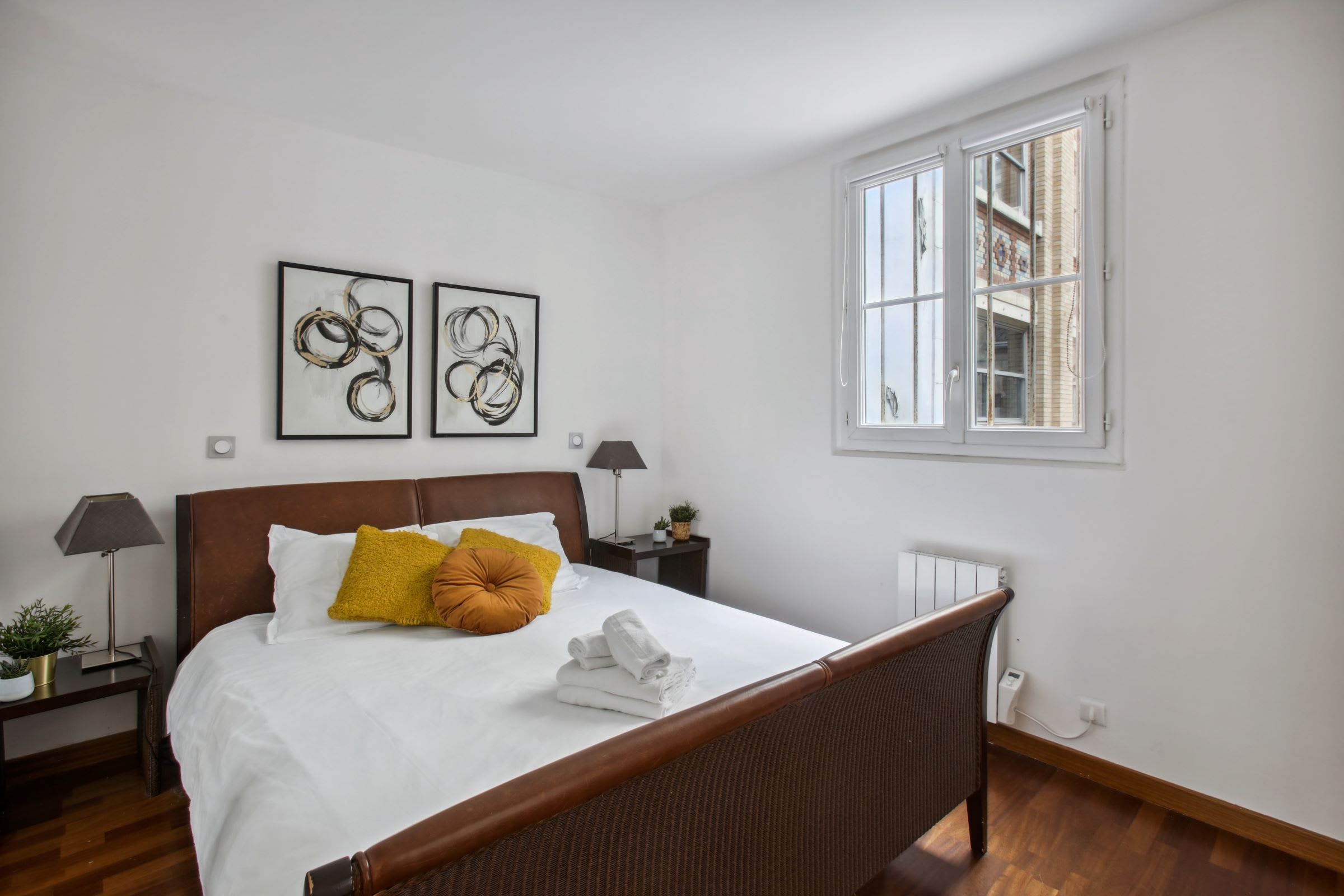 Un lit bien fait, avec du linge de lit blanc et une tête de lit marron, deux œuvres d’art abstraites encadrées accrochées au mur, une fenêtre donnant sur des immeubles et une petite table de chevet avec une lampe et une plante.