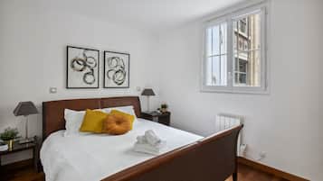 Un lit bien fait, avec du linge de lit blanc et une tête de lit marron, deux œuvres d’art abstraites encadrées accrochées au mur, une fenêtre donnant sur des immeubles et une petite table de chevet avec une lampe et une plante.