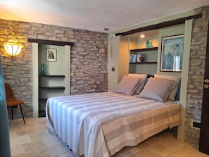 A bedroom with a stone wall, a bed with a striped bedspread, a nightstand, a chair, and a shelf with books and decorative items.