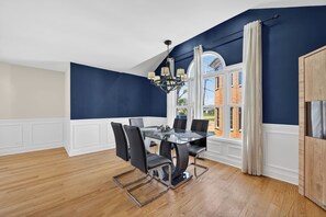 A dining room with a glass table and chairs, a chandelier, and a view of a brick building through large windows. - Huge 4BR Game Retreat · Sleeps 8 · Chicago (Glendale Heights)