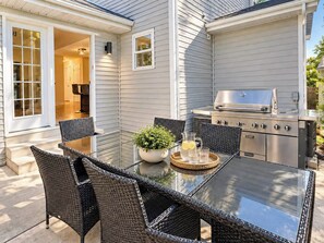 An outdoor patio with a glass-top table, wicker chairs, a grill, and a potted plant. - Huge 4BR Game Retreat · Sleeps 8 · Chicago (Glendale Heights)