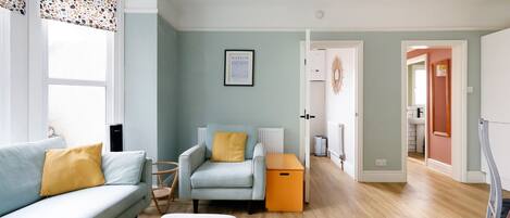 A living room with a sofa, armchair, ottoman, and a small table. There is a framed picture on the wall and a door leading to another room.