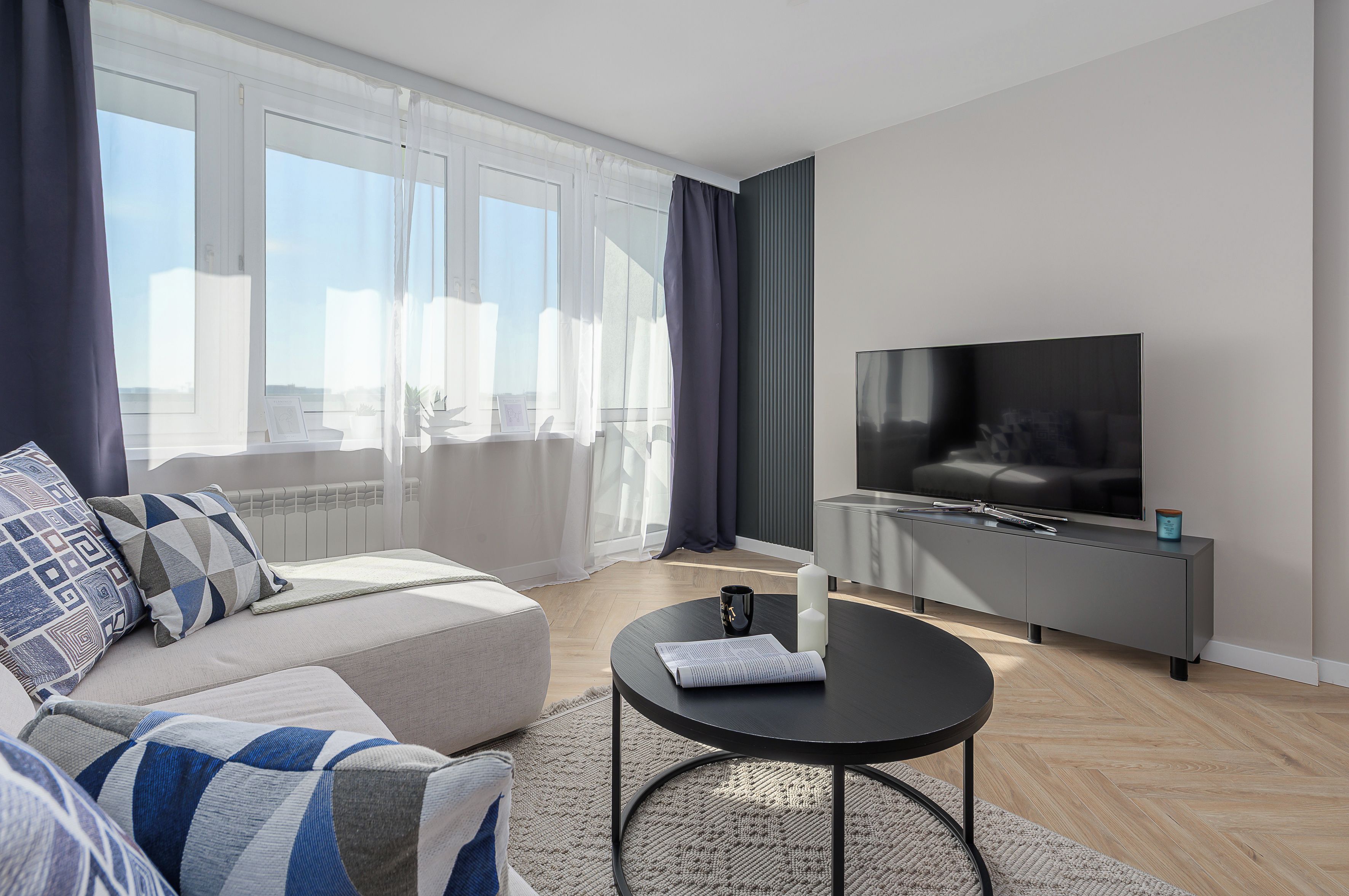 Een moderne woonkamer met een bank, salontafel, flatscreen televisie en een groot raam met gordijnen.