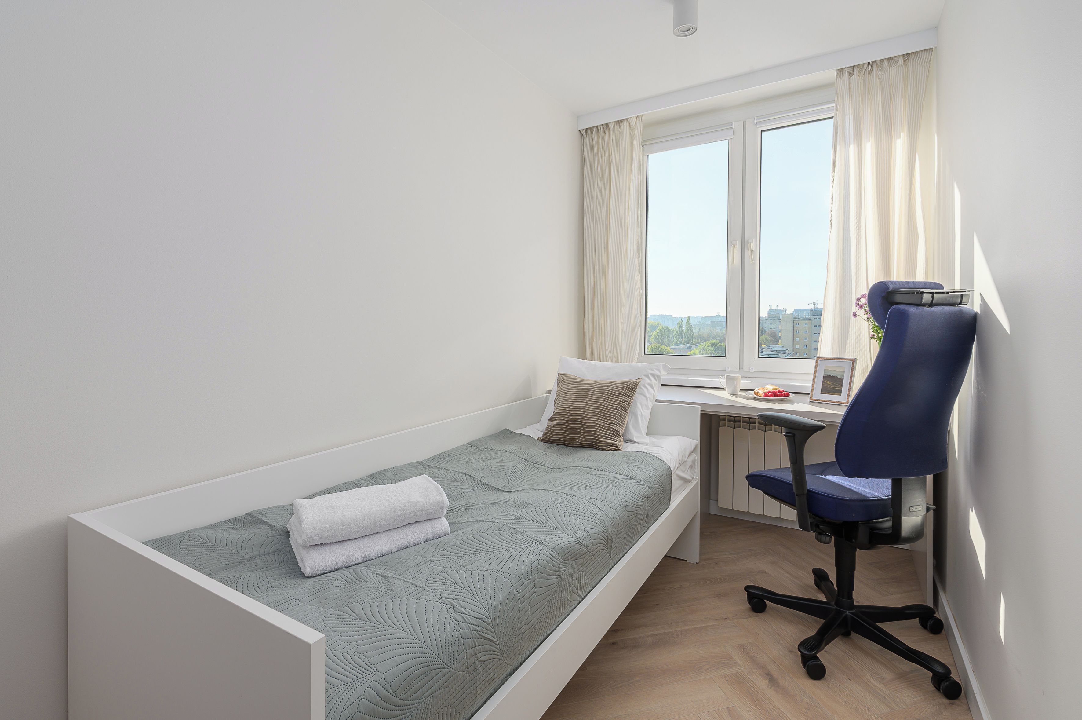 Una camera piccola con un letto singolo, una scrivania e una sedia, una finestra con tende e vista sugli edifici.