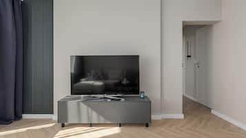 Un soggiorno moderno con una TV a schermo piatto su una console grigia, una finestra con persiane verticali e un pavimento in legno a spina di pesce.