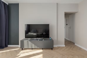 Een moderne woonkamer met een flatscreen televisie op een grijze console, een raam met verticale jaloezieën en een houten visgraatvloer.