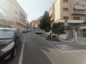 A street with parked cars and a scooter, buildings with balconies, and a gated entrance. - Aura Luxury Apartment: Sleeps 7, Garden, Parking, Near St. Peter's & Metro (Roma)