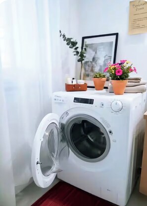 A front-loading washing machine with a control panel, a small brown storage basket, and two potted plants on top. - Aura Luxury Apartment: Sleeps 7, Garden, Parking, Near St. Peter's & Metro (Roma)