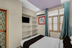 A bedroom with a bed, a TV mounted on the wall, a shelf unit, and a window with curtains.