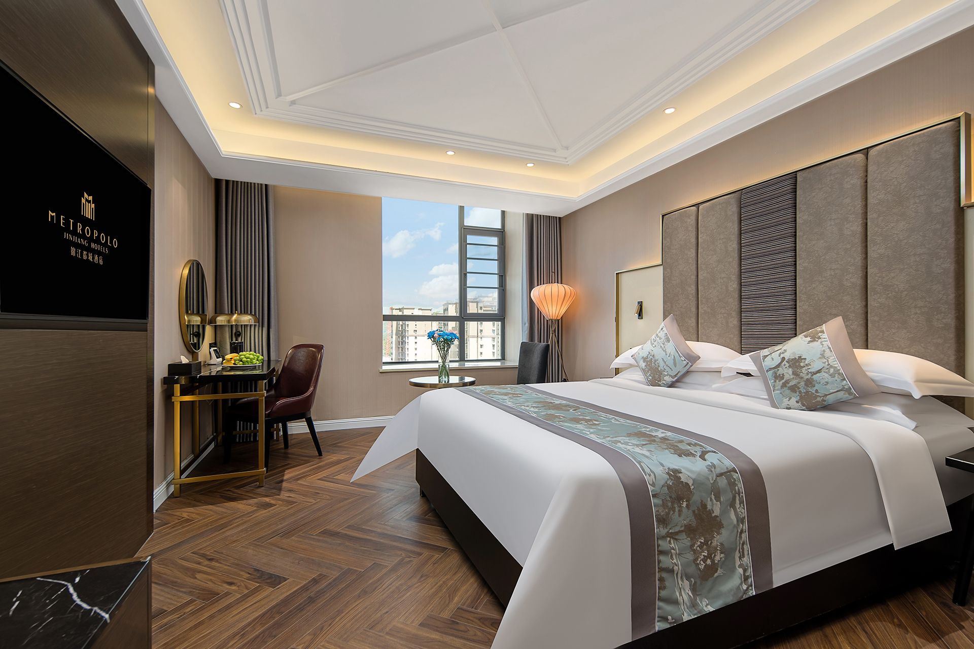 A hotel room with a large bed, a desk with a lamp, a chair, and a view of the city through a window.
