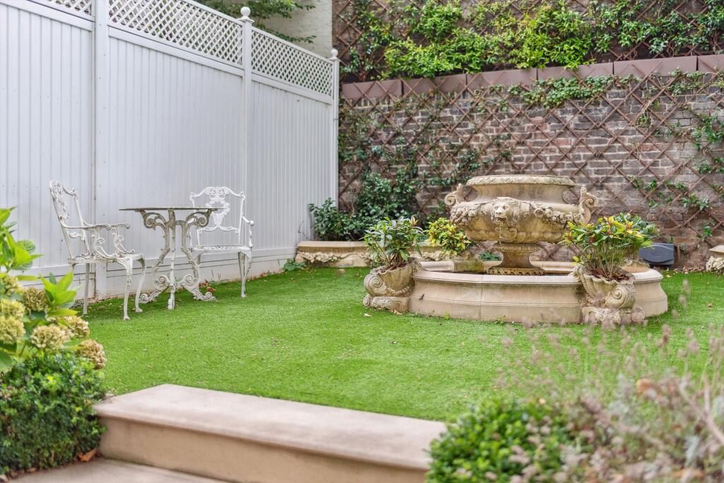 A garden with a fountain, white outdoor furniture, and a brick wall with climbing plants.