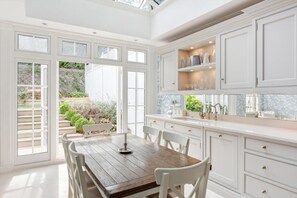 A bright kitchen with white cabinetry, a wooden dining table, and a view of a garden through large windows. - Distinguised 4-bedroom townhouse (South Kensington)