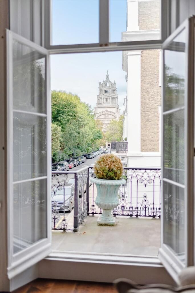 An open window with a view of a street, a building with a statue, and a potted plant on a balcony.