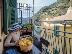 Ein Balkon mit Blick auf eine Küstenlandschaft, ein Korbwägelchen mit Zitronen und eine sitzende Person.