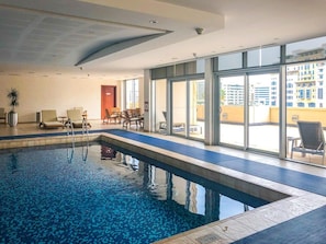 An indoor swimming pool with clear water, surrounded by lounge chairs and a glass wall offering a view of the city. - Vogue | Emaar Tower Al Rigga near Metro Station  (Dubai)