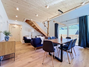 Ein geräumiges Zimmer mit Holzbalkendecke, einer Treppe, einem Essbereich mit Tisch und Stühlen sowie Blick durch große Fenster.