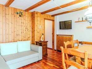 Ein Zimmer mit Holzwand, einem weißen Sofa, einem Holzesstisch mit Stühlen, einem an der Wand befestigten Fernseher und einem kleinen Schrank.