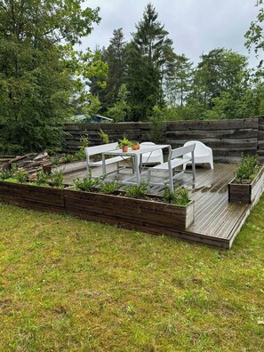 Una terraza de madera con una mesa y sillas, rodeada de bancales elevados y una valla de madera.