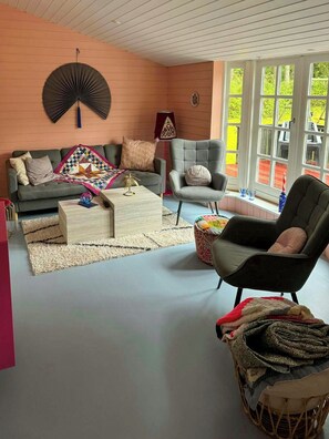 A living room with a sofa, armchair, coffee table, and a fan. There are windows with a view of greenery outside. - Summer house in pastel shades near Ebeltoft (Ebeltoft)