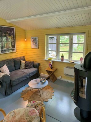A cozy living room with a sofa, a small round table, a decorative rug, a window with a view of greenery, and a wood-burning stove. - Summer house in pastel shades near Ebeltoft (Ebeltoft)