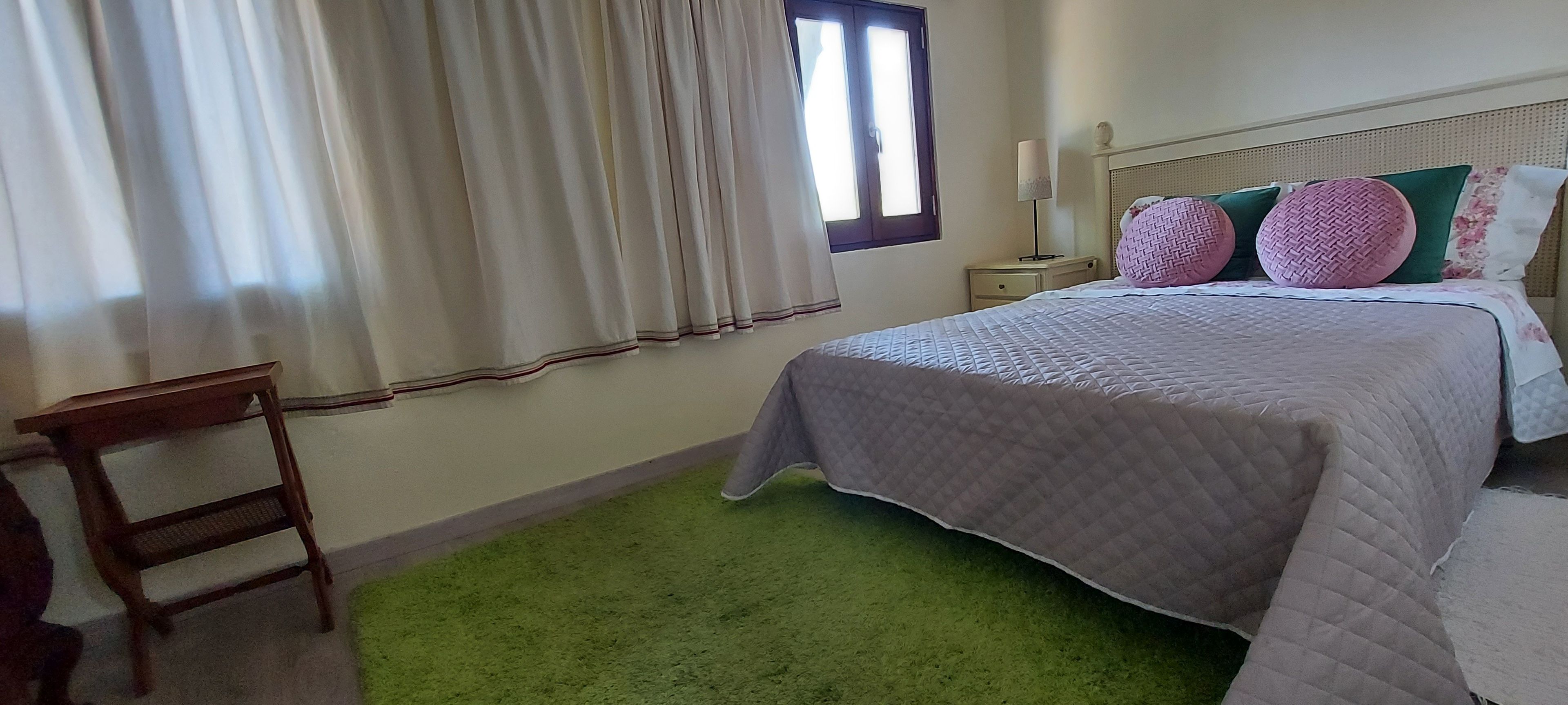 Een slaapkamer met een bed, een nachtkastje, een lamp, een stoel en een raam met gordijnen.