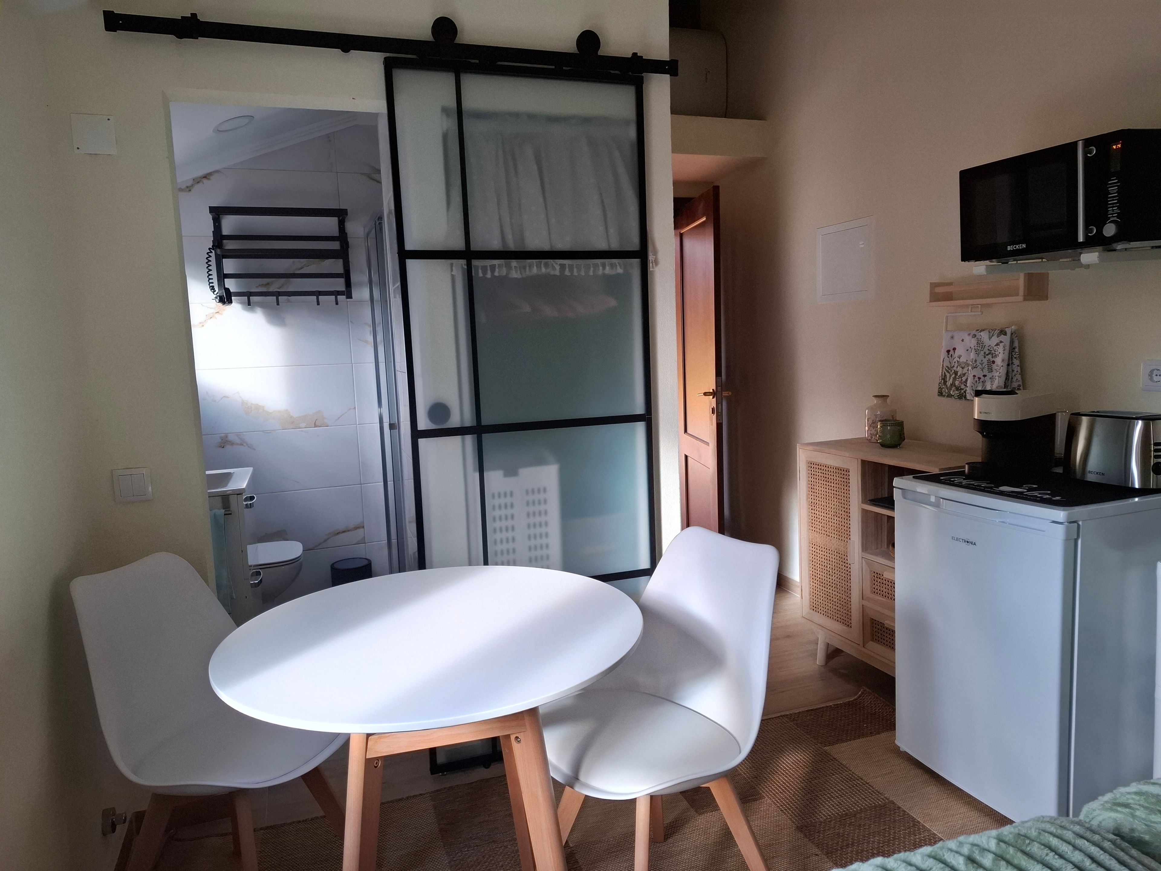 Een kleine keuken met een magnetron, een koffiezetapparaat en een koelkast. Er is een eethoek met een tafel en stoelen.