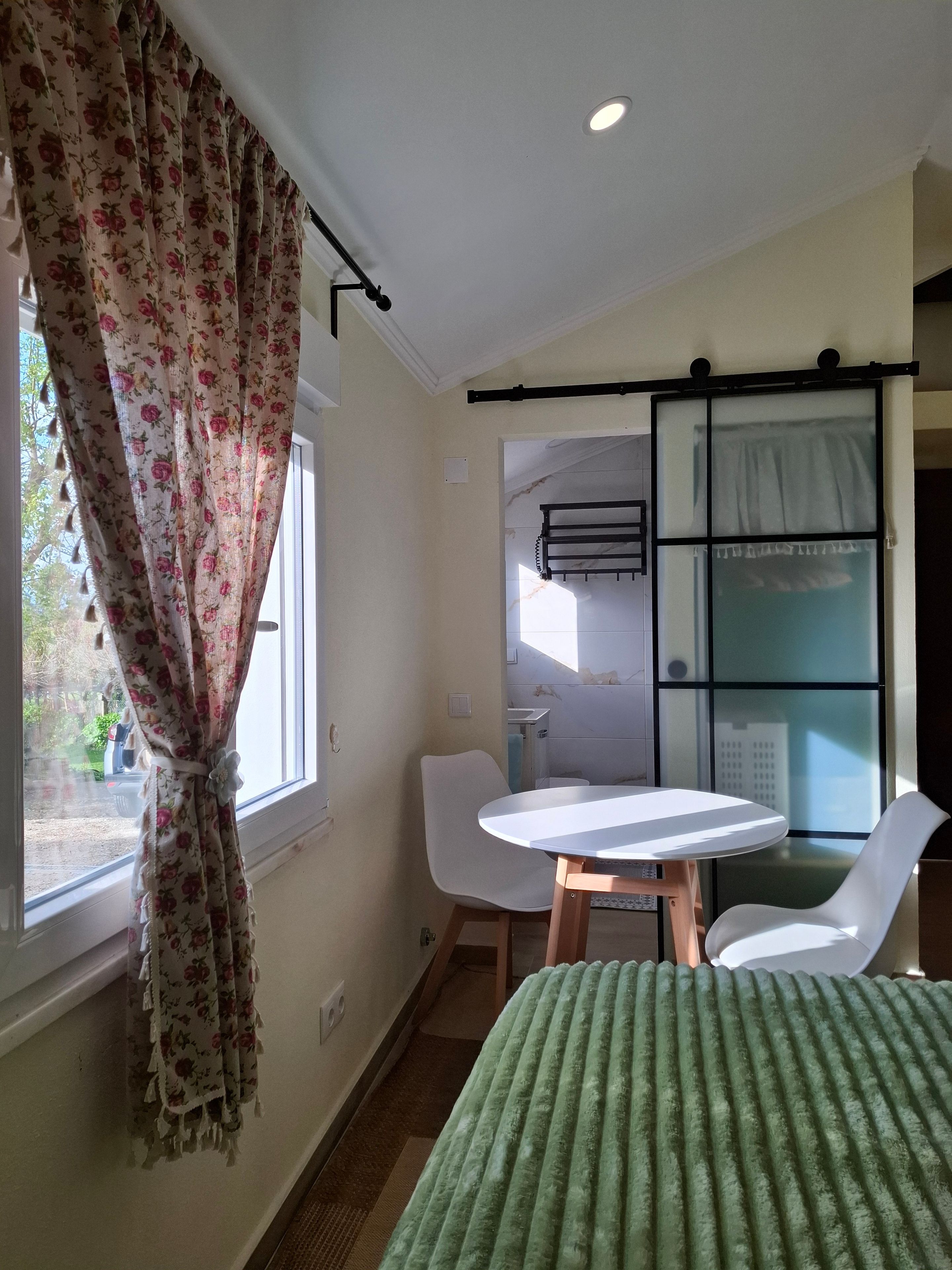 Ein Zimmer mit einem Bett, einem Tisch, zwei Stühlen, einem Fenster mit Vorhängen und einer Schiebetür.