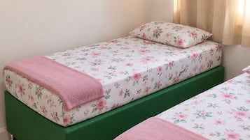 Un dormitorio con dos camas individuales, ropa de cama floral, un banco tapizado en verde y tres láminas botánicas enmarcadas en la pared.