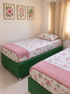 A bedroom with two single beds, floral bedding, a green upholstered bench, and three framed botanical prints on the wall.