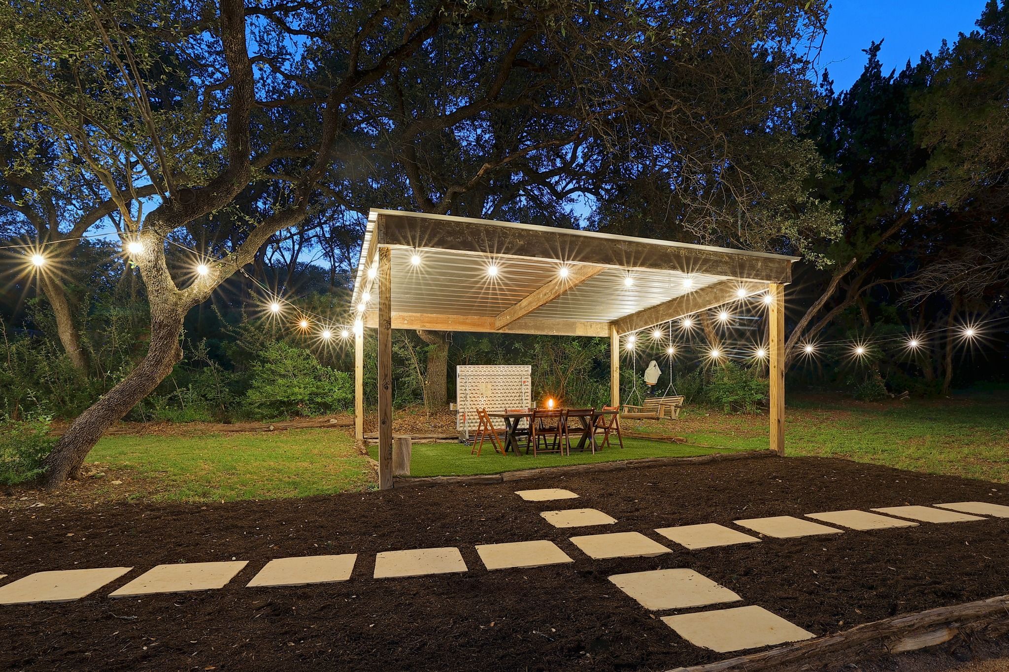 A covered outdoor seating area with a table and chairs, surrounded by a garden with stepping stones leading to it.
