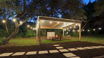 A covered outdoor seating area with a table and chairs, surrounded by a garden with stepping stones leading to it.