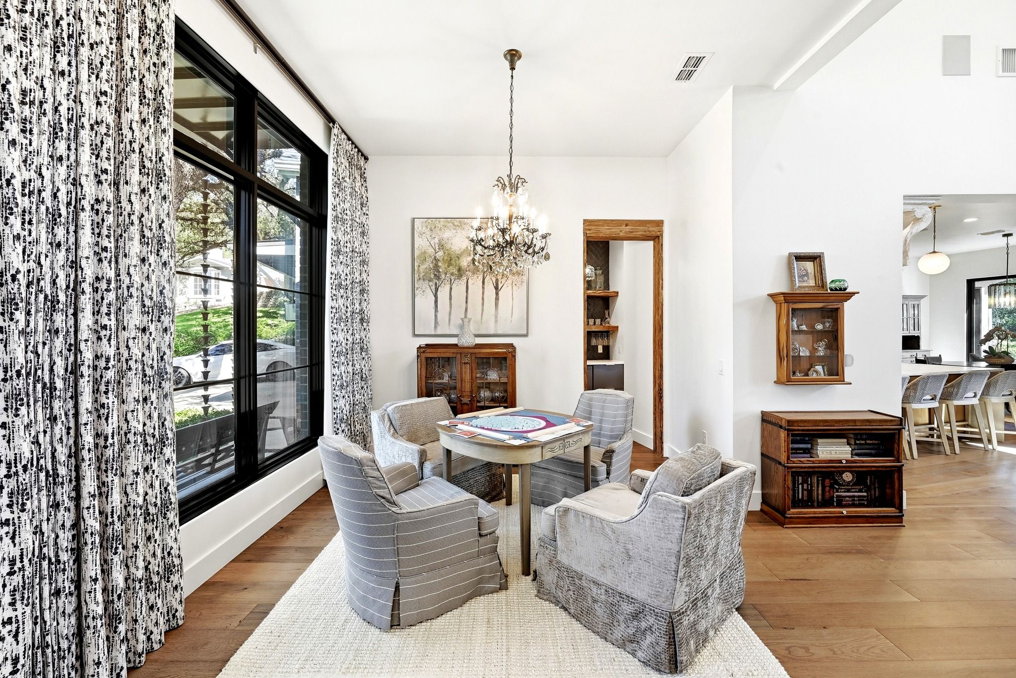 A well-lit living room with a large window, a chandelier, and a dining area with a round table and chairs.