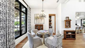 A well-lit living room with a large window, a chandelier, and a dining area with a round table and chairs.